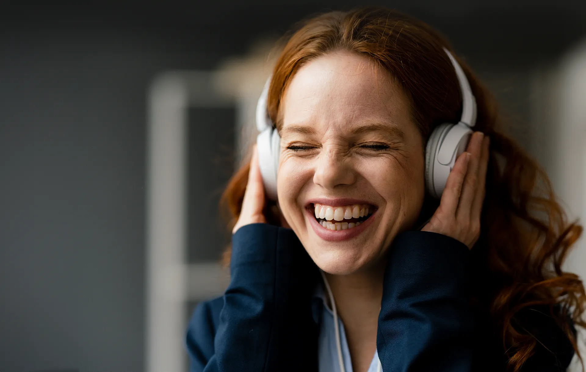 Portrait einer lachenden Businessfrau mit rötlichen Haaren, welche die Hände über weiße Kopfhörer hält und Musik hört